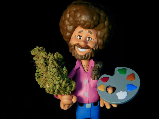 A humorous Bob Ross-style figurine holding a large cannabis bud and a painter's palette against a black background, symbolizing creativity and marijuana culture.