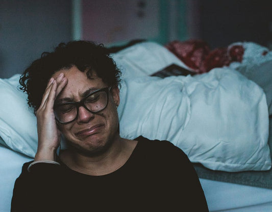 A woman with glasses sits on the floor beside a bed, holding her head and crying in emotional distress after a sleepless night. The dim lighting and unmade bed in the background create an exhausted, overwhelmed mood.