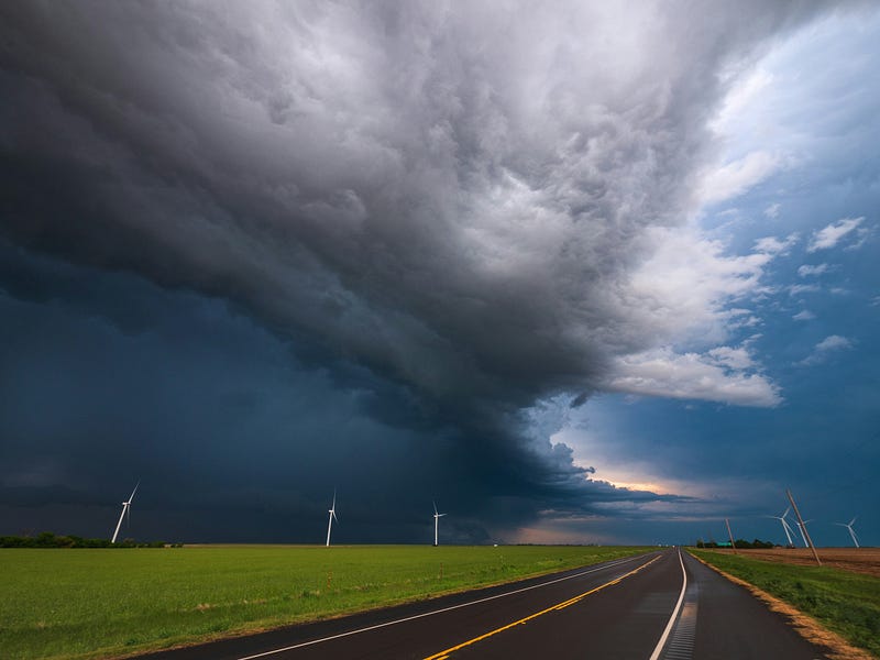 Storm clouds roll over a rural highway cutting through open green fields, with wind turbines standing in the distance as a dark, heavy storm front collides with lighter sky on the horizon.