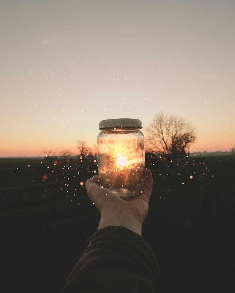 A hand holding a clear glass jar glowing with warm light at sunset, with a dark field and bare trees in the background.