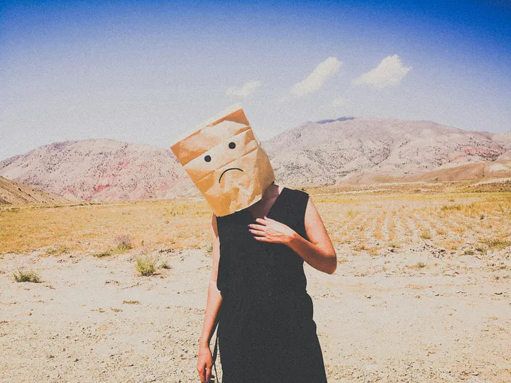 A person standing in a barren landscape with a paper bag covering their face, symbolizing emotional disconnection and loss of identity during PMDD dysphoria.