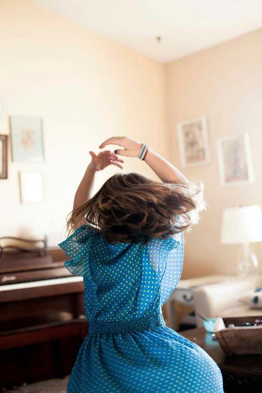 Person dancing freely indoors with arms raised and hair in motion, wearing a blue polka-dot dress in a softly lit living room, conveying joy, movement, and emotional release.
