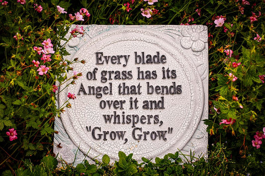 Stone plaque in a garden surrounded by flowers with the quote "Every blade of grass has its Angel that bends over it and whispers, Grow, Grow"