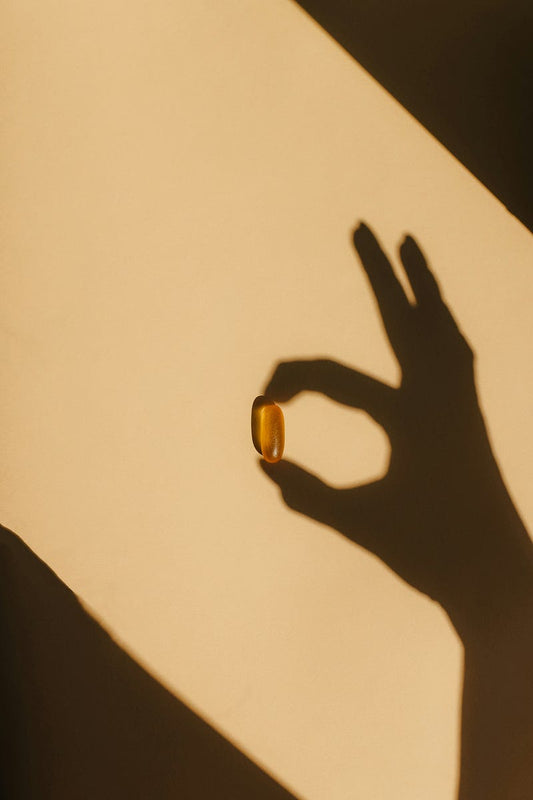 A hand’s shadow holds a single amber pill between two fingers against a warm, golden background.