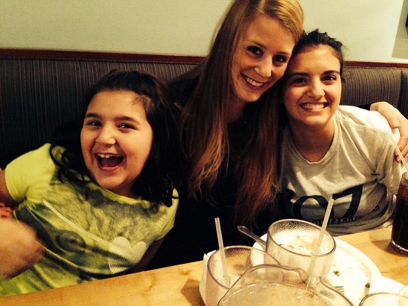 Three smiling girls sitting together in a restaurant booth, leaning close for a photo with milkshake glasses on the table.