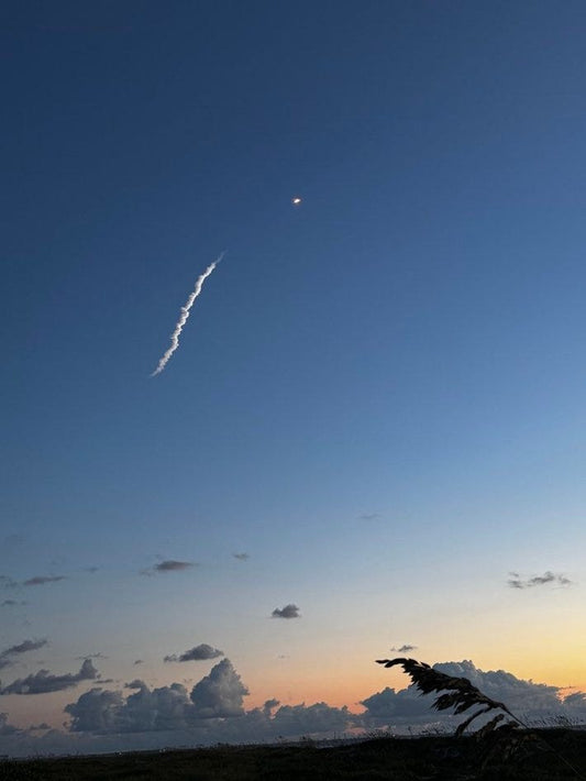 Distant rocket glowing in a twilight sky above coastal dunes and sea oats along Florida's Space Coast, with sunset colors on the horizon.