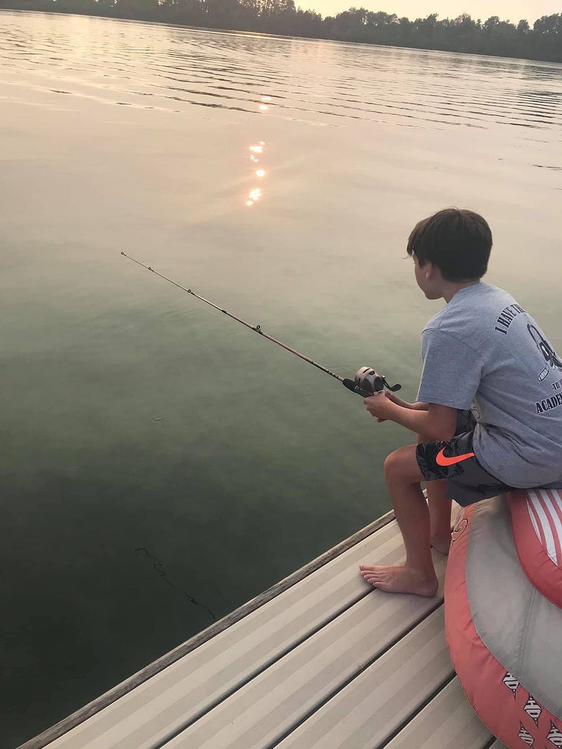 A young boy sits on the edge of a dock at sunset, barefoot and focused as he fishes on a calm lake, with soft sunlight reflecting on the water and trees lining the horizon.