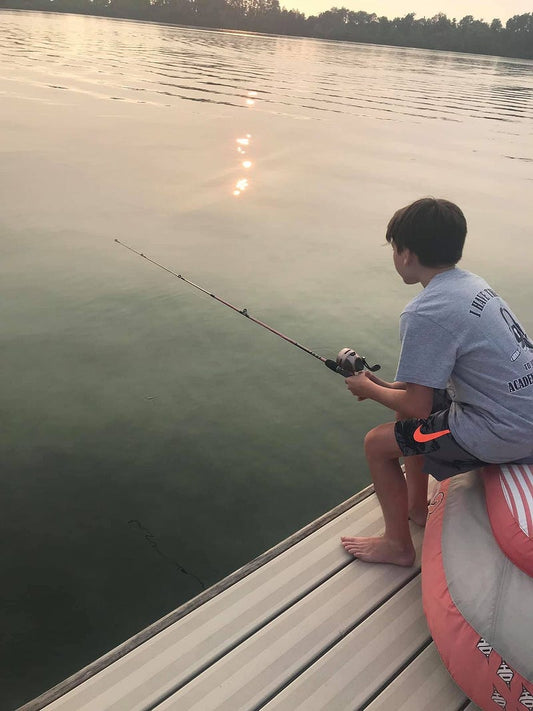 A young boy sits on the edge of a dock at sunset, barefoot and focused as he fishes on a calm lake, with soft sunlight reflecting on the water and trees lining the horizon.