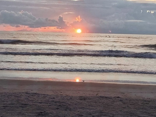 Sunrise over the Atlantic Ocean at Cocoa Beach, with the sun glowing orange on the horizon, soft waves rolling toward shore, and clouds lit in pink and gold above the water.