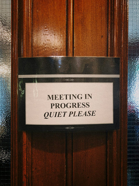 A sign reading "Meeting in Progress - Quiet Please" hangs on a wooden door with textured glass panels, marking the entrance to a private meeting room.
