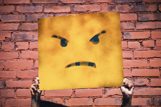 A yellow sign with an angry cartoon face is held up against a red brick wall. Only the holder's hands are visible.