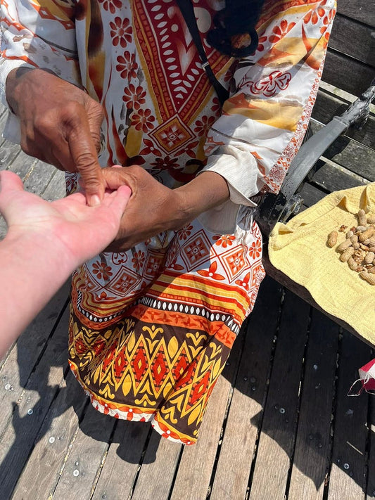 A woman in a patterned orange, yellow, and white dress holds another person's hand palm-to-palm while standing on the Atlantic City boardwalk. A small pile of peanuts rests on a towel beside her.