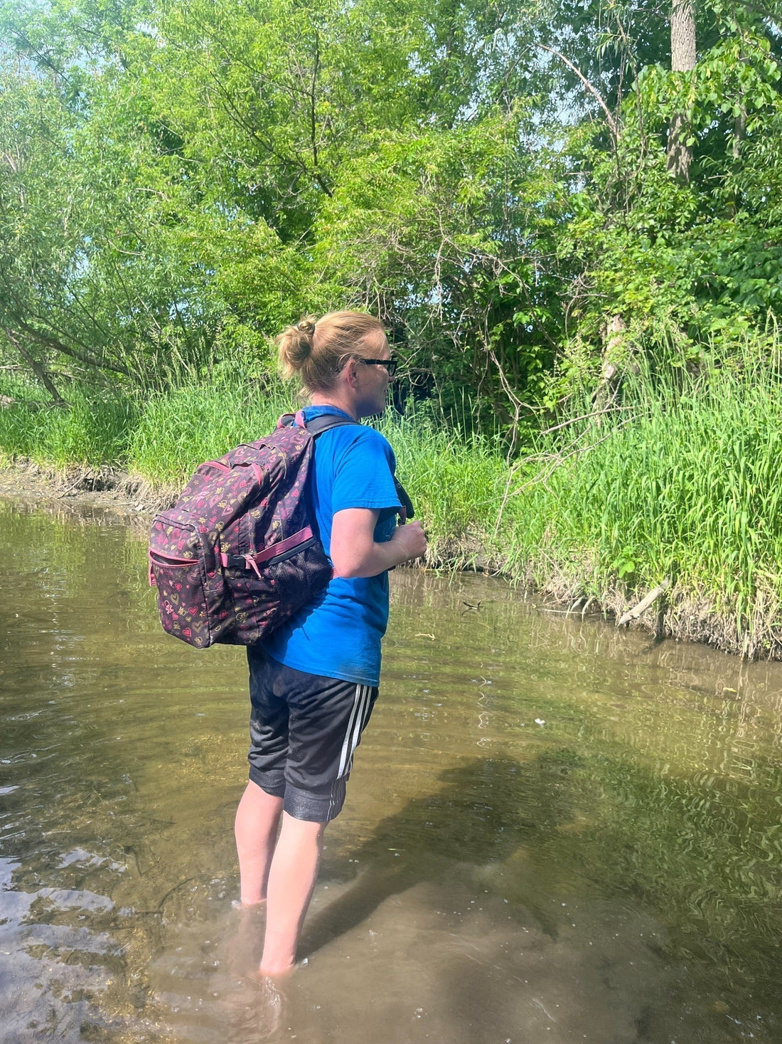 A woman with a backpack stands barefoot in a shallow creek surrounded by green trees and tall grasses.