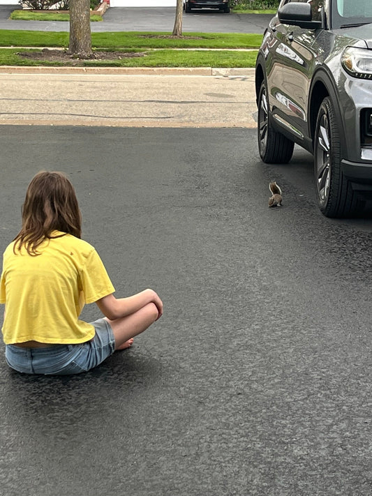 A young girl sits on a rain-soaked driveway studying a baby squirrel with a crooked foot, watching its every move like a wildlife researcher conducting an important field study.