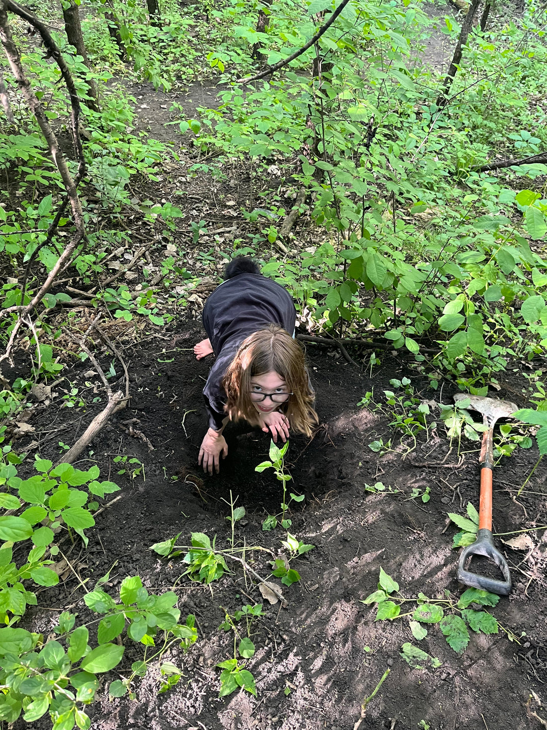 A girl with glasses digs a deep hole in the woods, surrounded by green forest plants, with a shovel resting nearby.