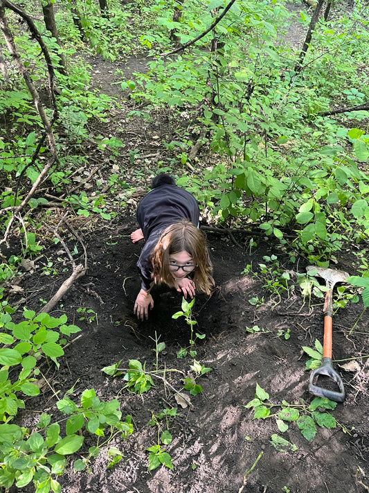 A girl with glasses digs a deep hole in the woods, surrounded by green forest plants, with a shovel resting nearby.