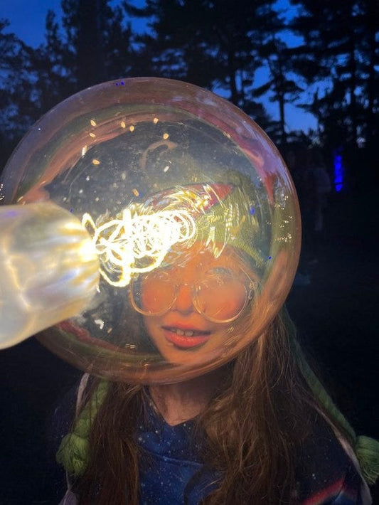 A young girl wearing large round glasses looks through a giant glowing bubble at night.
