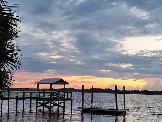 A wooden dock sits over calm water at sunset in St. Augustine, the sky glowing pink and orange beneath heavy clouds. The stillness of the scene feels peaceful and grounding.