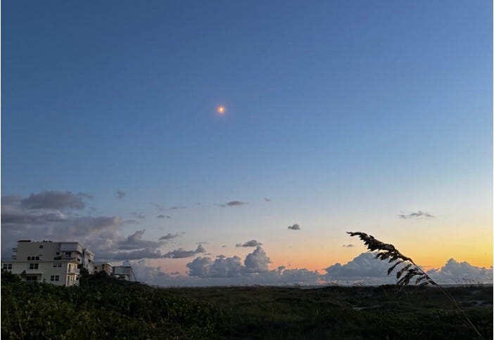 Distant rocket glowing in a twilight sky above coastal dunes and sea oats along Florida's Space Coast, with sunset colors on the horizon.