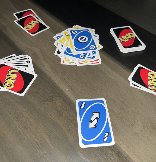 Uno cards spread across a wooden table during a game, with a blue skip card in the center and several small piles of cards around it, suggesting an active, slightly chaotic round of play.