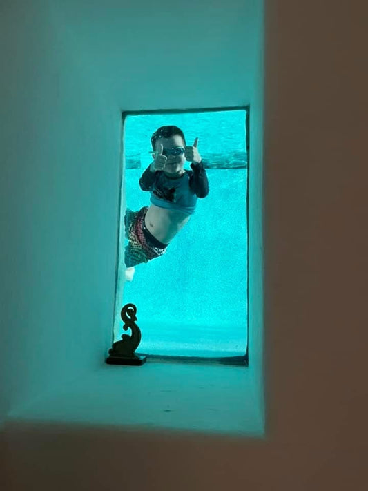 Young boy with goggles on giving thumbs up under water while looking at camera through a window in the pool.